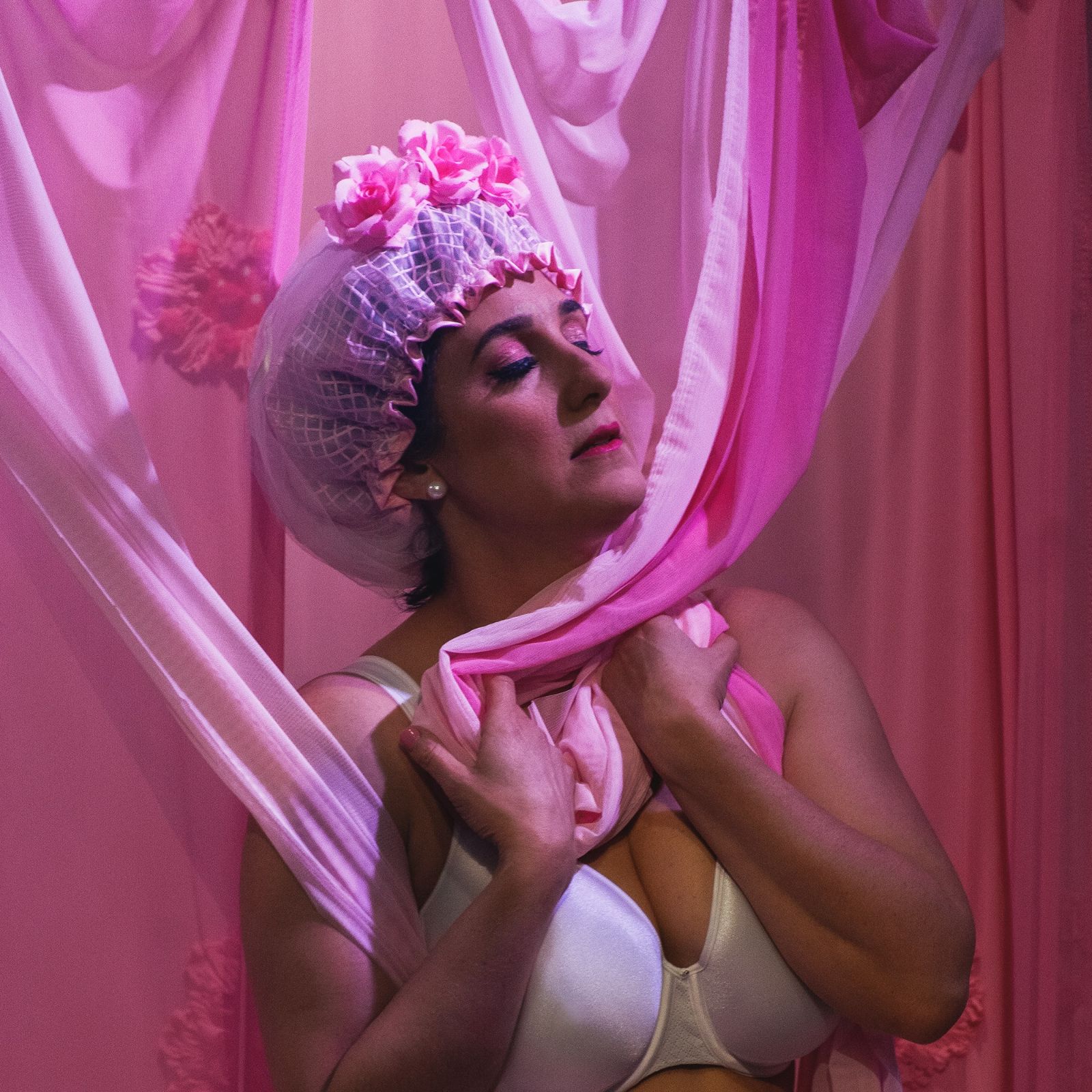 Woman in white bra and pink bonnet with loofas on top drapes pink curtains aroun