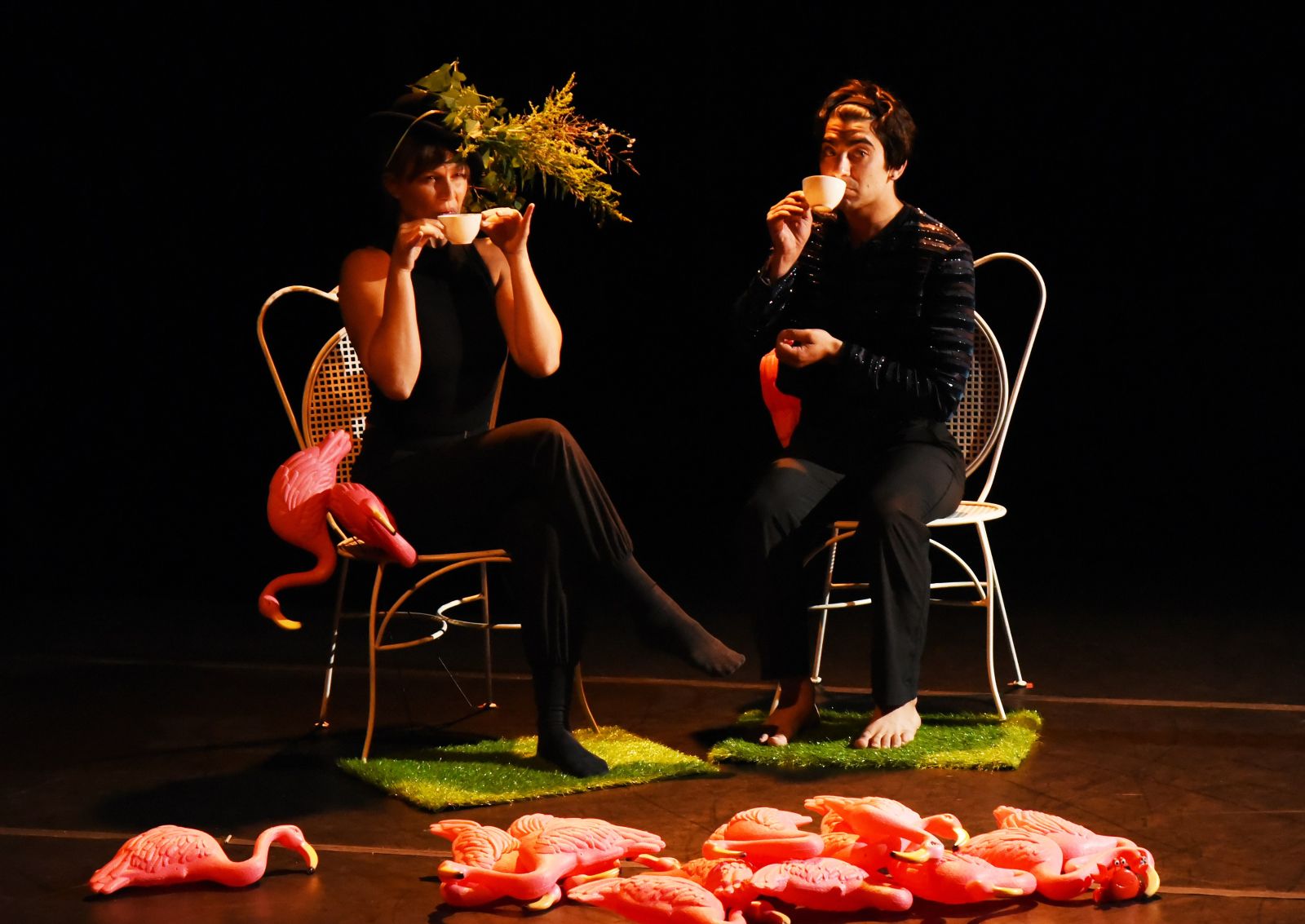 Two dancers in black seated in lawn chairs with plastic flamingos on the floor