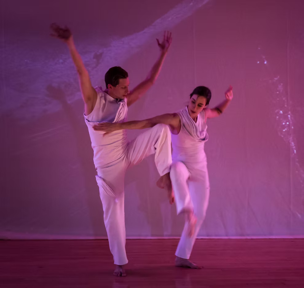 Two dancers in white posing with their legs connected against a pink background